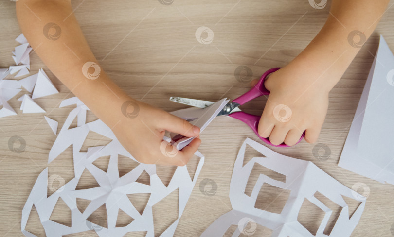 Скачать Ребенок создает дома за столом белые бумажные снежинки. Ребенок вырезает ножницами узоры на бумаге. Украшение для зимнего праздника. фотосток Ozero