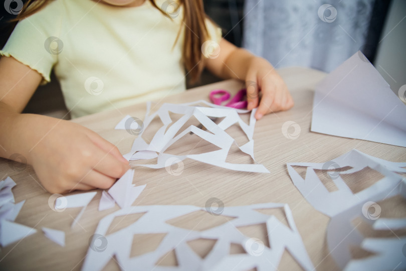 Скачать Ребенок создает дома за столом белые бумажные снежинки. Ребенок вырезает ножницами узоры на бумаге. Украшение для зимнего праздника. фотосток Ozero