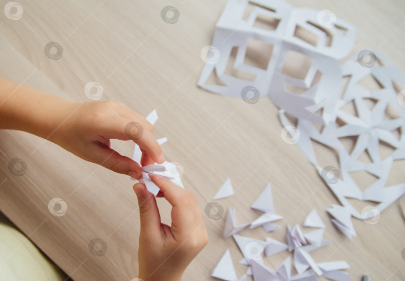 Скачать Ребенок создает дома за столом белые бумажные снежинки. Ребенок вырезает ножницами узоры на бумаге. Украшение для зимнего праздника. фотосток Ozero