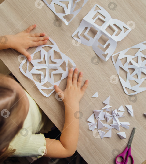 Скачать Ребенок создает дома за столом белые бумажные снежинки. Ребенок вырезает ножницами узоры на бумаге. Украшение для зимнего праздника. фотосток Ozero