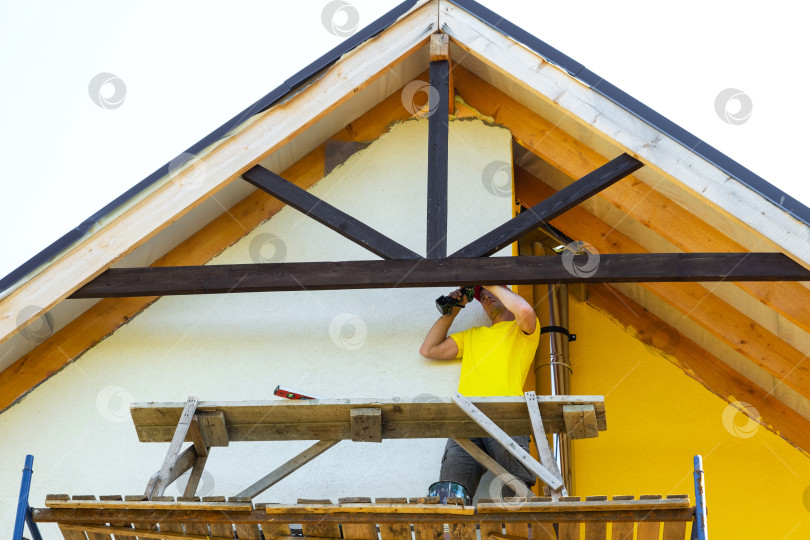 Скачать Профессиональный строитель крепит новую балку к высокому фронтону, обеспечивает видимую изоляцию и сайдинг, внешнюю отделку дома, имеет под рукой инструменты и средства безопасности. фотосток Ozero