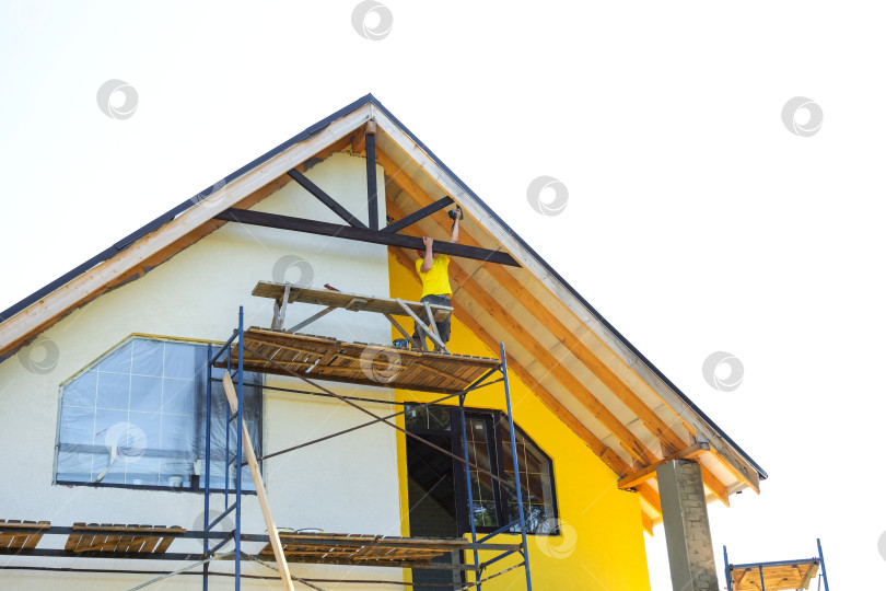 Скачать Рабочий на многоуровневых строительных лесах устанавливает отделку фасада желто-белого дома, создавая современный экстерьер, командную работу и концепцию проекта реконструкции. фотосток Ozero