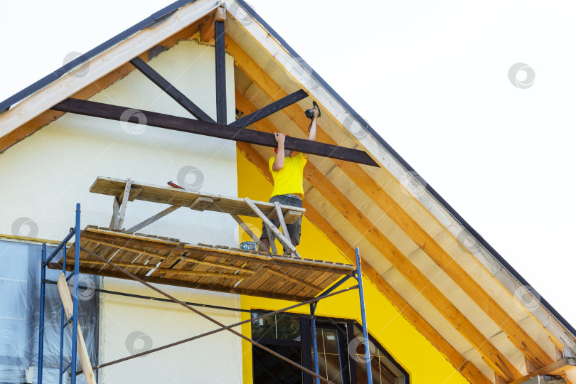 Скачать Мужчина поднимается по лестнице, чтобы добраться до фронтона, подготавливает деревянные панели к монтажу, желтая жилая застройка, сделай сам, безопасность на высоте. фотосток Ozero