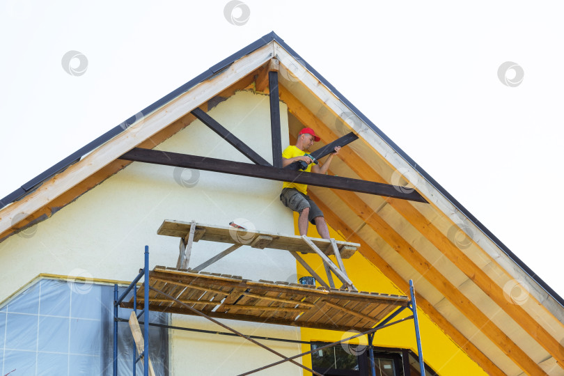 Скачать Плотник в каске красит декоративную перекладину фронтона со строительных лесов, видны инструменты и оборудование, современный дом желтого цвета, внимание к безопасности. фотосток Ozero