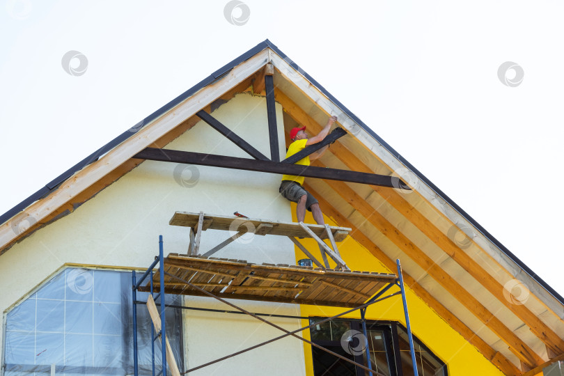 Скачать Профессионал в желтой рубашке заделывает швы в декоративной деревянной отделке, фасаде фронтона дома, рабочей зоне, на виду у всех шпаклевочный пистолет и защитная каска. фотосток Ozero