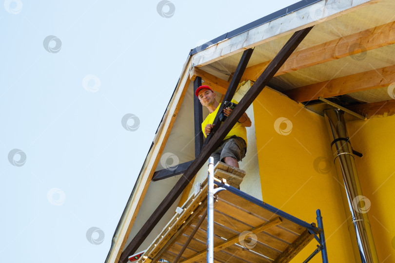 Скачать Рабочий сидит на строительных лесах, осматривая законченный декор деревянного фронтона, современный желтый дом, внешнюю архитектуру, акцент на достижениях и безопасности. фотосток Ozero