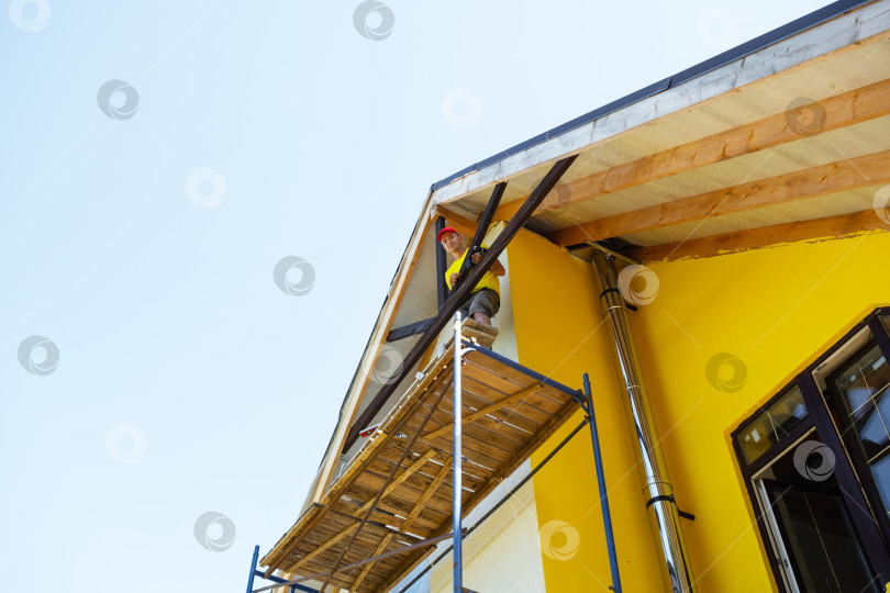 Скачать Строитель на верхней платформе завершает отделку фронтона, бело-желтый дом, свет заката, последний этап строительства дома, видны ручные инструменты. фотосток Ozero