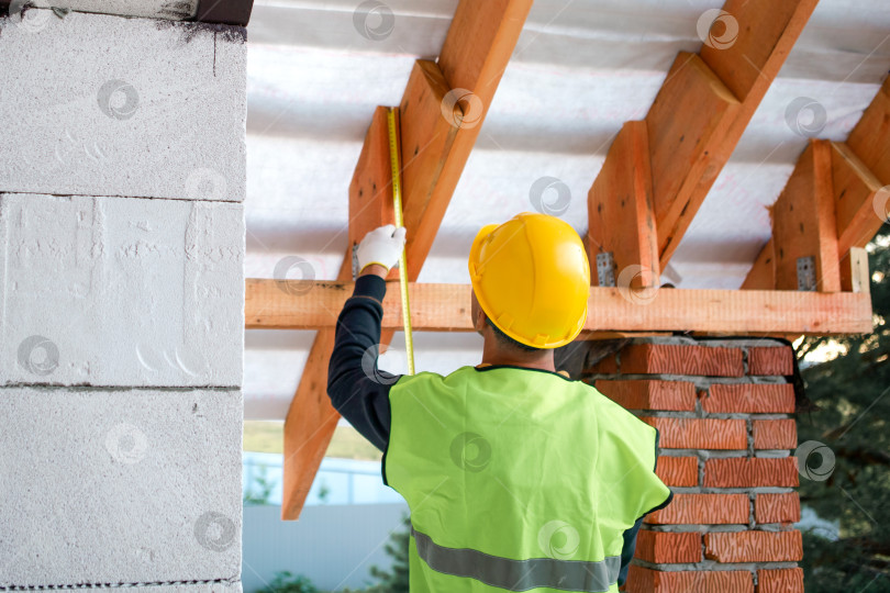 Скачать Мужчина-строитель в жилете проверяет выравнивание стропил, установку болтов для крепления каркаса крыши, видимую кирпичную опору и изоляцию, строительство дома. фотосток Ozero