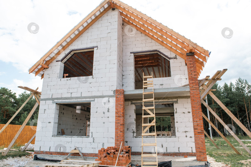 Скачать Строящийся современный каменный дом, видимые деревянные стропила и балки крыши, активная сборка каркаса крыши завершают этап строительства. фотосток Ozero
