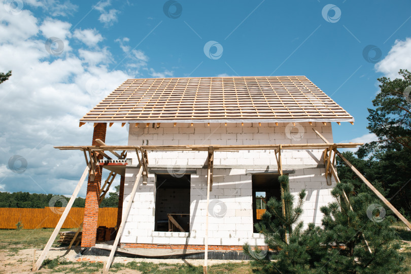 Скачать Вид спереди на коттедж в евростиле с деревянными лагами на крыше, незавершенная система стропил, яркое небо, современная фаза строительства. фотосток Ozero