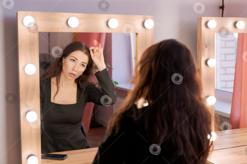 Скачать Женщина смотрит на свое отражение в зеркале для макияжа, и свет касается ее волос. фотосток Ozero
