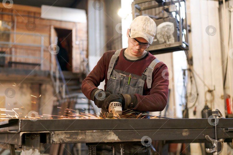 Скачать Молодой рабочий шлифует металлическую раму с искрами в мастерской фотосток Ozero