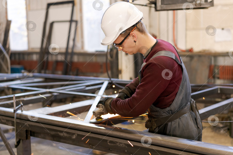 Скачать Рабочий-подмастерье шлифует большую стальную раму в мастерской фотосток Ozero