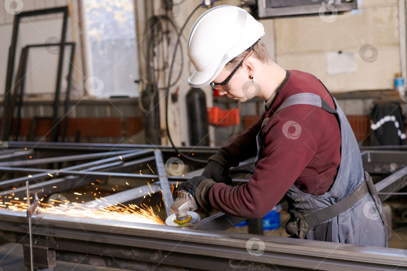 Скачать Молодой рабочий в каске с искрами шлифует стальную раму фотосток Ozero