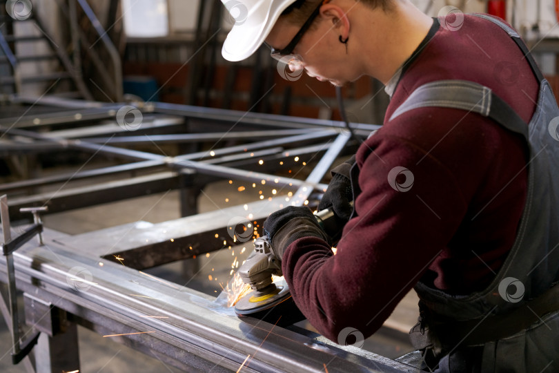 Скачать Молодой рабочий в белой каске шлифует стальные конструкции фотосток Ozero