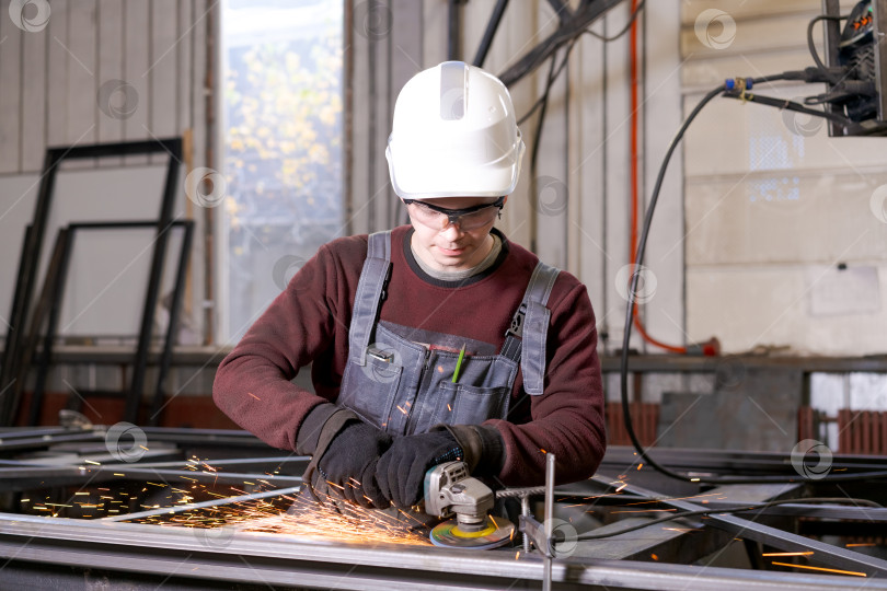 Скачать Рабочий по металлу в белой каске шлифует стальную раму фотосток Ozero