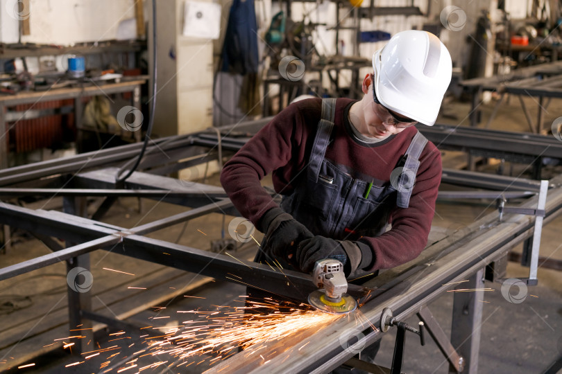 Скачать Молодой рабочий в каске шлифует стальную раму на заводе фотосток Ozero