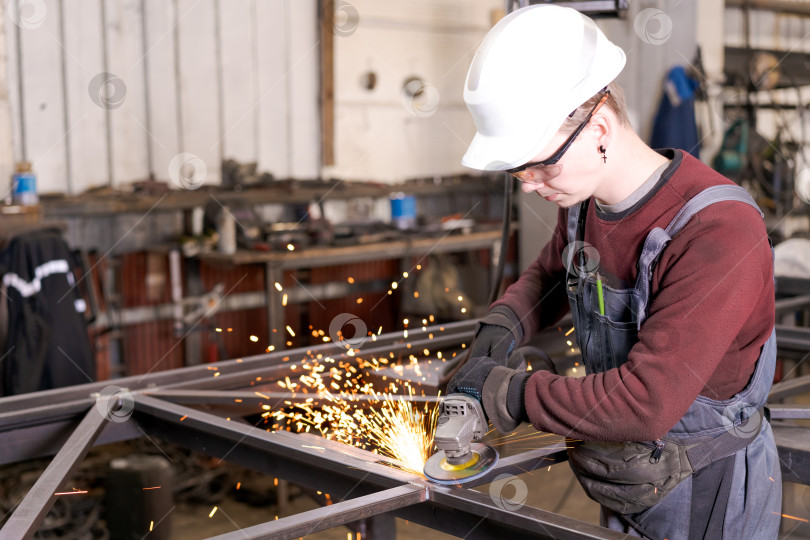 Скачать Молодой подмастерье в белой каске шлифует металлическую раму фотосток Ozero