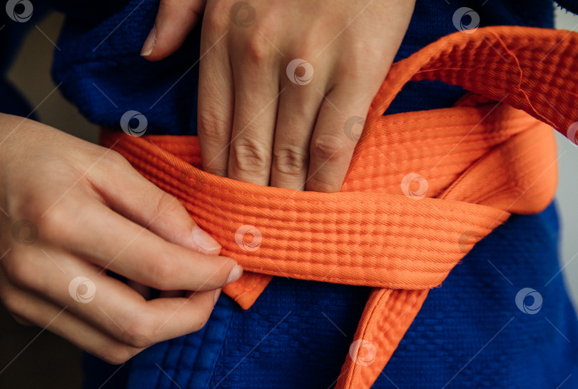 Скачать Деталь синего спортивного костюма для дзюдо, перевязанная оранжевым поясом, демонстрирующая тренировки по боевым искусствам. Правила завязывания пояса. фотосток Ozero