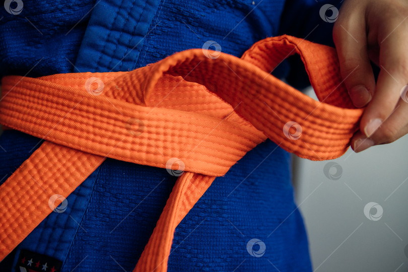 Скачать Деталь синего спортивного костюма для дзюдо, перевязанная оранжевым поясом, демонстрирующая тренировки по боевым искусствам. Правила завязывания пояса. фотосток Ozero