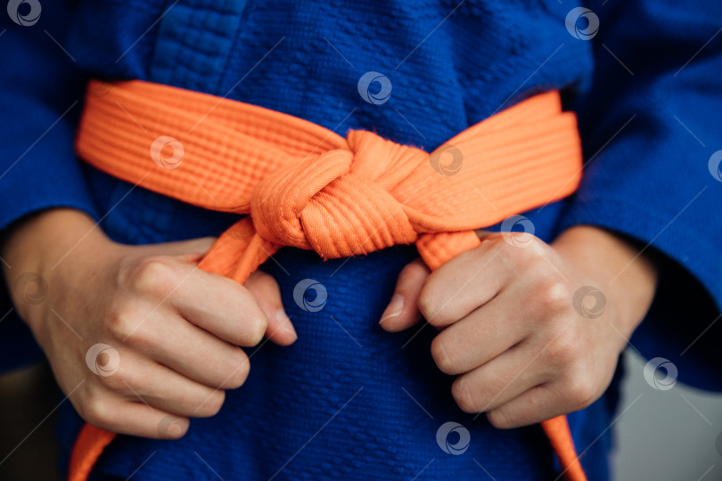 Скачать Деталь синего спортивного костюма для дзюдо, перевязанная оранжевым поясом, демонстрирующая тренировки по боевым искусствам. Правила завязывания пояса. фотосток Ozero