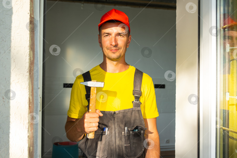 Скачать Портрет строителя в красной шапочке с молотком в руках, готового к плотницким работам, забиванию гвоздей и ремонту дома, мастер на все руки, набор инструментов на месте. фотосток Ozero