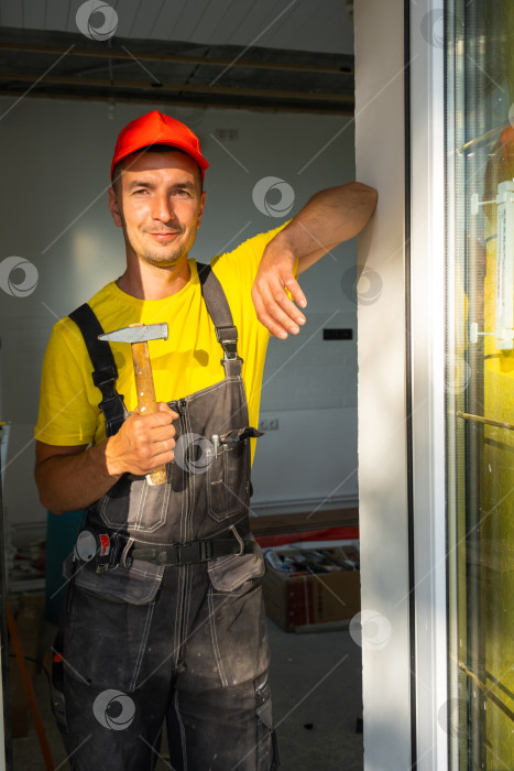 Скачать Молодой строитель стоит у окна с молотком в руках, готовый к строительству дома, ремонту, сборке мебели, услугам разнорабочего, визиту квалифицированного специалиста. фотосток Ozero
