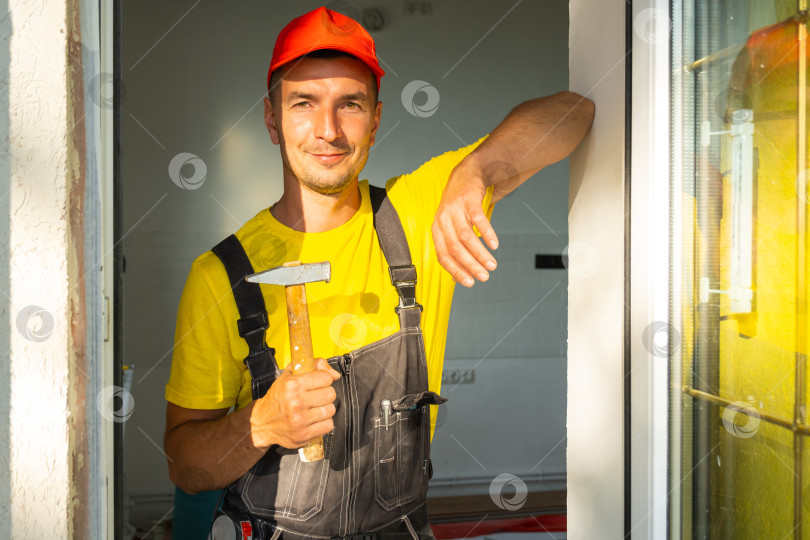 Скачать Специалист по ремонту, держащий молоток у окна, готов к забиванию гвоздей, обрамлению, столярным работам, забиванию молотком, установке на дому, услугам разнорабочего и знанию инструментов. фотосток Ozero