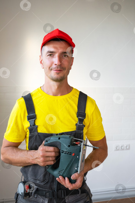 Скачать Ремонтник в красной шапочке с аккумуляторной дрелью в руках, фасад дома, универсальные инструменты для ремонта, услуги разнорабочего, работа на дому, техническое обслуживание и монтаж зданий. фотосток Ozero