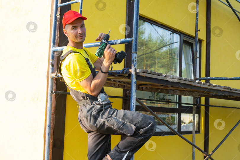 Скачать Строитель, стоящий на строительных лесах с электроинструментом, реставрация фасадов, покраска и монтаж, услуги разнорабочего на дому, профессиональный ремонт и консультации. фотосток Ozero
