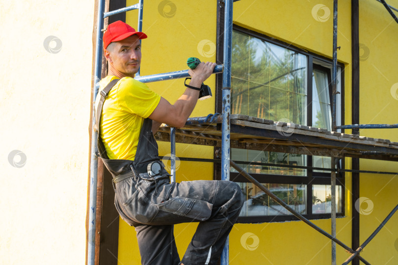 Скачать Разнорабочий на строительных лесах с дрелью, ремонт фасадов, профессиональные наружные работы, реновация, строительные инструменты, выездной специалист по благоустройству и покраске дома. фотосток Ozero