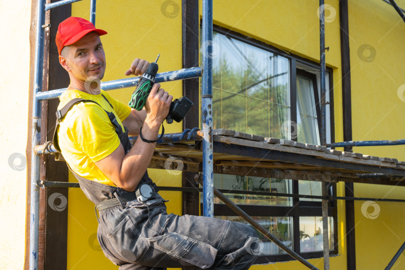 Скачать Строитель в красной шапочке с отверткой на строительных лесах, фасад дома, ремонт дома, набор инструментов, мастер на все руки, приезжающий для домашнего ремесла, технического обслуживания и усовершенствования. фотосток Ozero