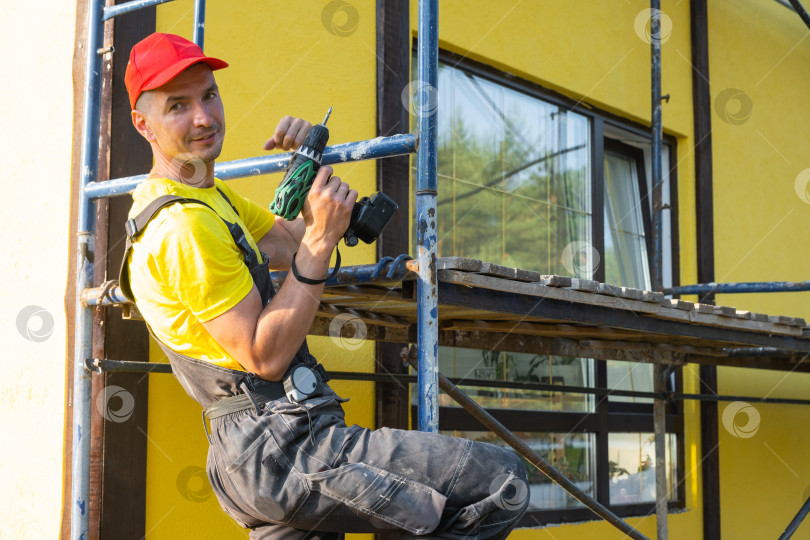Скачать Рабочий-ремонтник с дрелью на строительных лесах, фасад дома, обслуживание на дому, оборудование для строительства, покраска, техническое обслуживание, разнорабочий по ремонту, специалист по отделке. фотосток Ozero