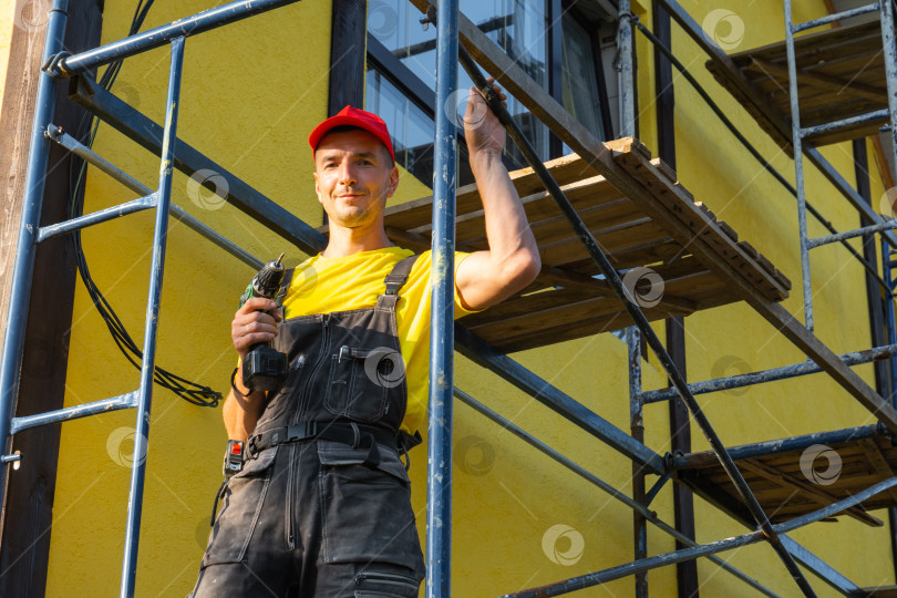 Скачать Строитель с малярным валиком, строительными лесами, готовый к наружным стеновым работам, благоустройству дома, инструментом мастера на все руки, посещению сервисной службы, профессиональному строительному ремонту. фотосток Ozero