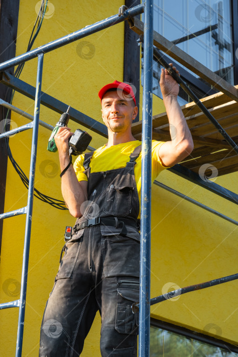 Скачать Ремонтник в спецодежде, на строительных лесах с малярным валиком, покраска фасада, консультации по внешнему ремонту дома, сервис на месте, набор инструментов для разнорабочего. фотосток Ozero
