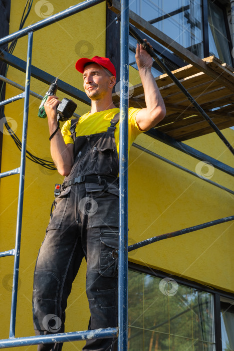 Скачать Юный строитель с малярным валиком, стоящий на строительных лесах, покраска фасадов, наружные работы, инструменты разнорабочего, выезд на дом для ремонта, техническое обслуживание. фотосток Ozero