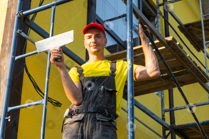 Скачать Улыбающийся мастер-ремонтник с роликом на строительных лесах, покраска дома, техническое обслуживание здания, вызов мастера на все руки, эксперт-консультант по наружным работам. фотосток Ozero