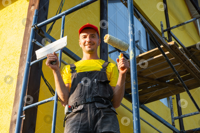 Скачать Мастер на все руки с малярным валиком в руках, стоящий на строительных лесах, наружная покраска дома, текущий ремонт, посещение сервисной службы на дому, набор инструментов, профессиональная работа. фотосток Ozero