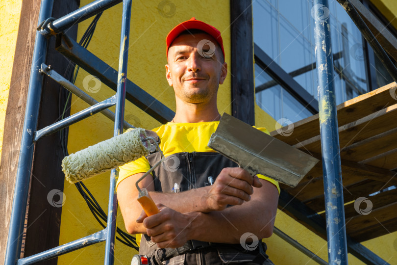 Скачать Портрет молодого строителя с большим малярным валиком, ремонт фасада, строительные леса, техническое обслуживание, выезд на дом, полное обслуживание разнорабочего, инструменты для ремонта. фотосток Ozero
