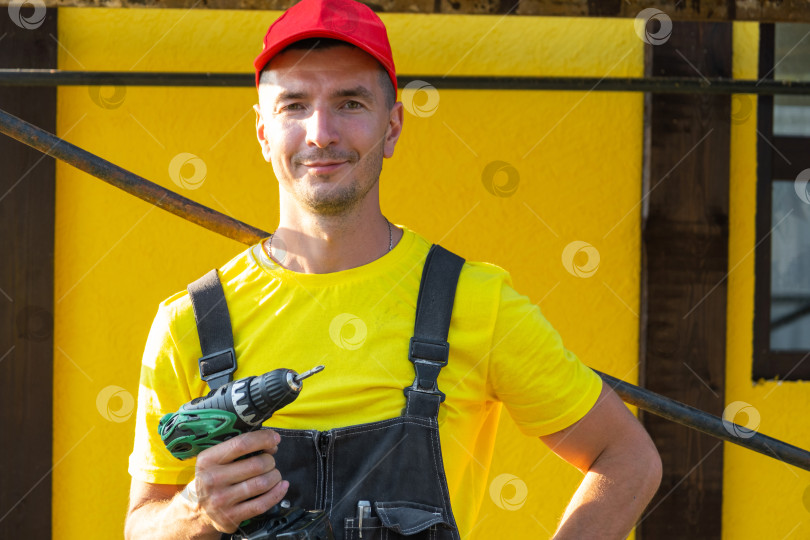 Скачать Улыбающийся молодой строитель в желтой рубашке с дрелью в руках, домашний ремонт своими руками, строительные работы, оборудование для разнорабочих, частный подрядчик, набор инструментов, визит специалиста. фотосток Ozero
