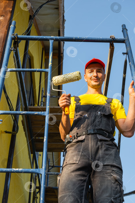 Скачать Молодой строитель с роликом в руках, строительные леса, покраска фасадов, ремонт стен, обустройство дома, инструменты для разнорабочего, ремонтный бизнес, посещение консультационной службы. фотосток Ozero