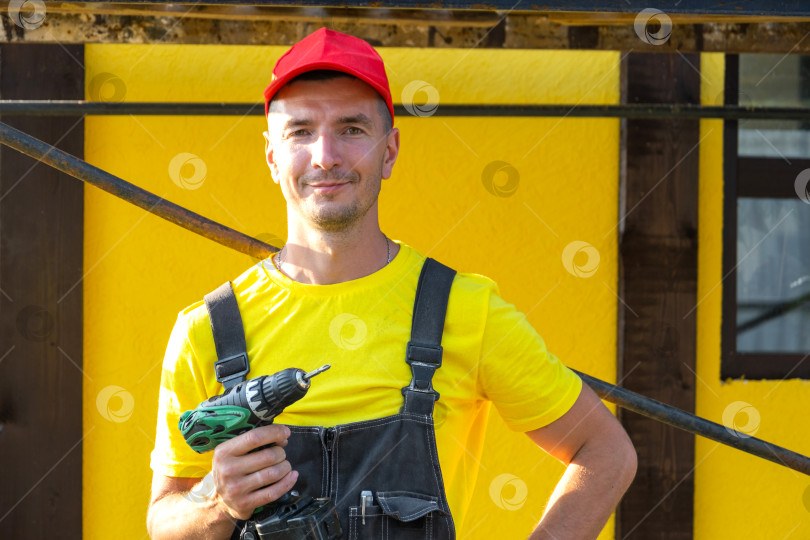 Скачать Портрет строителя с дрелью в руках, стоящий возле строящегося дома, услуги по ремонту, частный бизнес разнорабочего, инструменты, навыки обустройства дома своими руками. фотосток Ozero