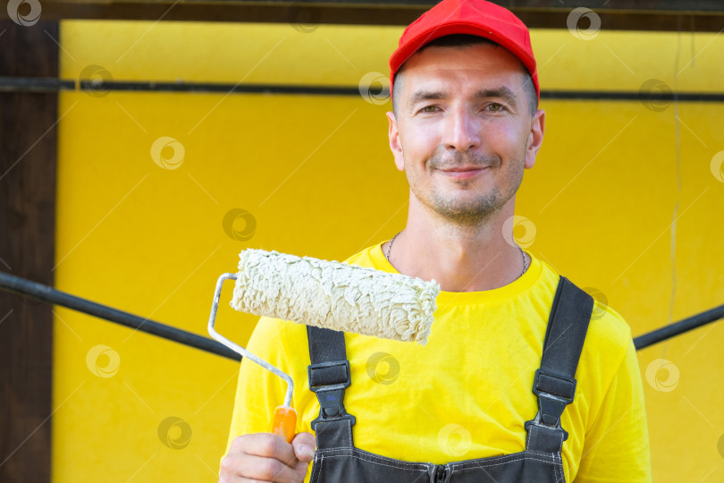 Скачать Строитель с малярным валиком в руках, стоящий перед домом, покраска стен и обустройство дома, услуги частного мастера на все руки, инструменты для ремонта на дому. фотосток Ozero