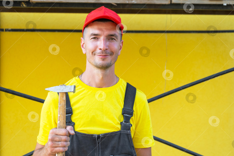 Скачать Молодой мастер на все руки в красной шапочке с молотком в руках, строительство дома, плотницкие работы, забивание гвоздей, ремонтные работы, частный бизнес, услуги своими руками, выезд на место, мастерство. фотосток Ozero