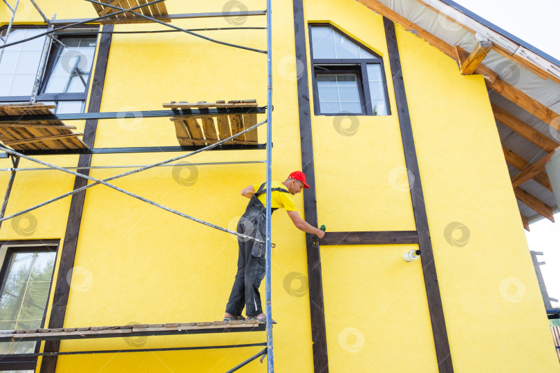 Скачать Рабочий на строительных лесах, устанавливающий декоративную древесину для фасада фахверкового дома, отделка фасадов домов, профессиональный мастер, занимающийся облицовкой домов. фотосток Ozero