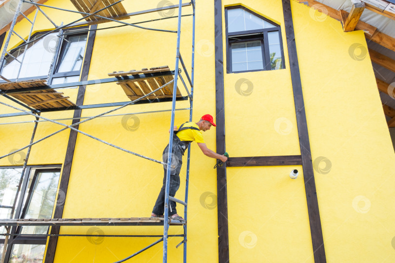 Скачать Мастер прикрепляет вертикальные деревянные декоративные элементы к фасаду, стиль фахверк, внешняя отделка дома, благоустройство здания, работа по частному проекту. фотосток Ozero