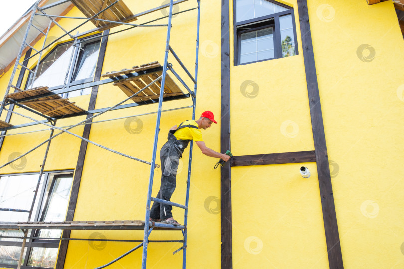 Скачать Фасадный рабочий, крепящий деревянные рейки для декоративной отделки в стиле фахверк, стильный внешний вид дома, работа мастера, элементы облицовки, услуги по ремонту. фотосток Ozero