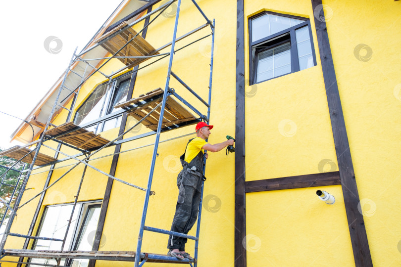 Скачать Мастер в красной шапочке устанавливает декоративную древесину на высоком фасаде, выполняет внешнюю отделку дома, строительство из фахверка, улучшает облицовку, монтажные работы на высоте. фотосток Ozero