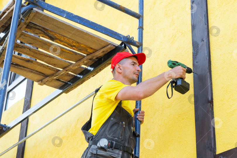 Скачать Рабочий использует отвертку для крепления деревянной отделки фахверкового фасада, декоративных деталей экстерьера, обустройства дома на месте, частного ремонтного проекта. фотосток Ozero