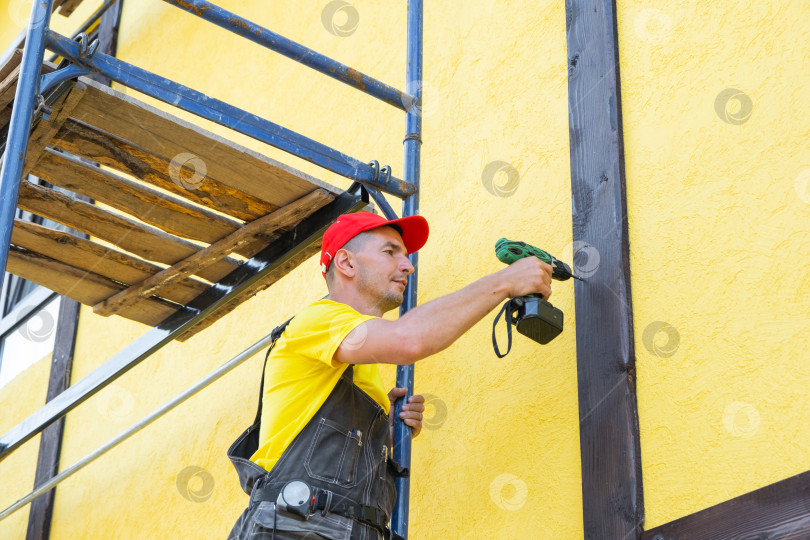 Скачать Мастер монтирует деревянные элементы на стену дома, дизайн в стиле фахверк, декоративную облицовку, улучшение внешнего вида дома своими руками, ремонтные работы в частном порядке. фотосток Ozero